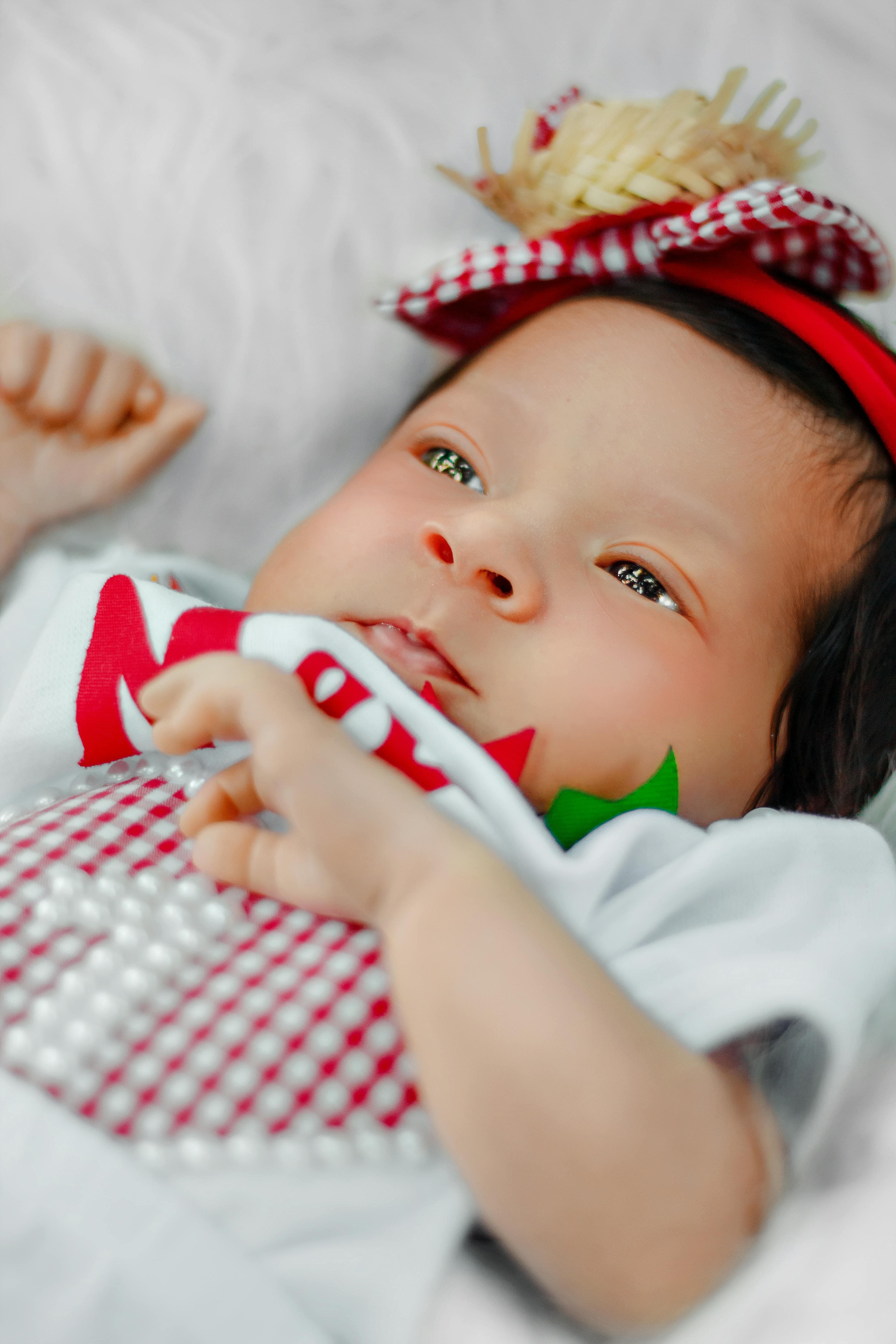 Un bebé acostado sobre una superficie blanca, con una diadema roja de cuadros y un pequeño adorno de paja, vestido con ropa blanca y roja, mirando hacia un lado.