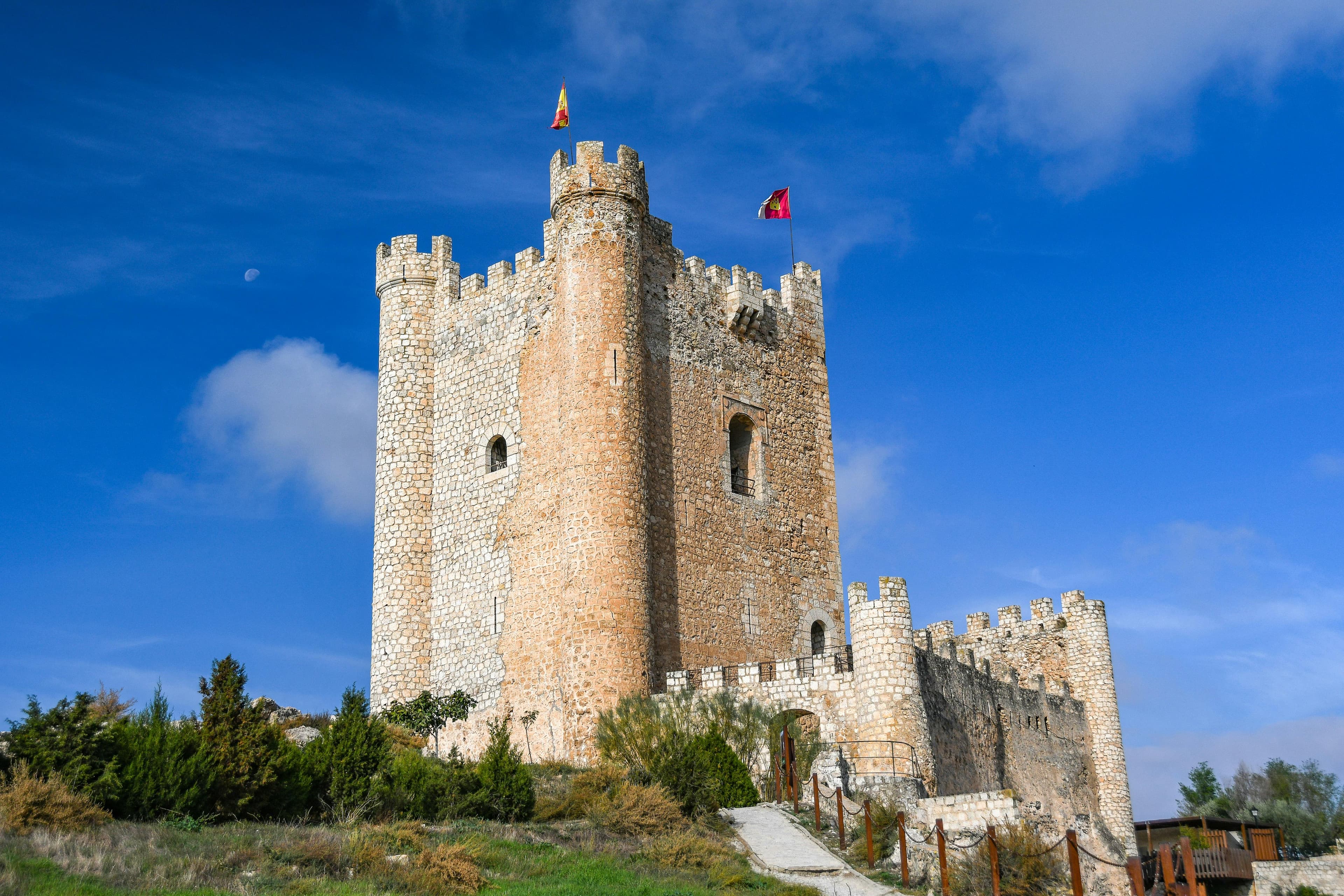 Castillo medieval de piedra con torres almenadas, situado en una colina, con banderas ondeando en la cima bajo un cielo azul con algunas nubes.