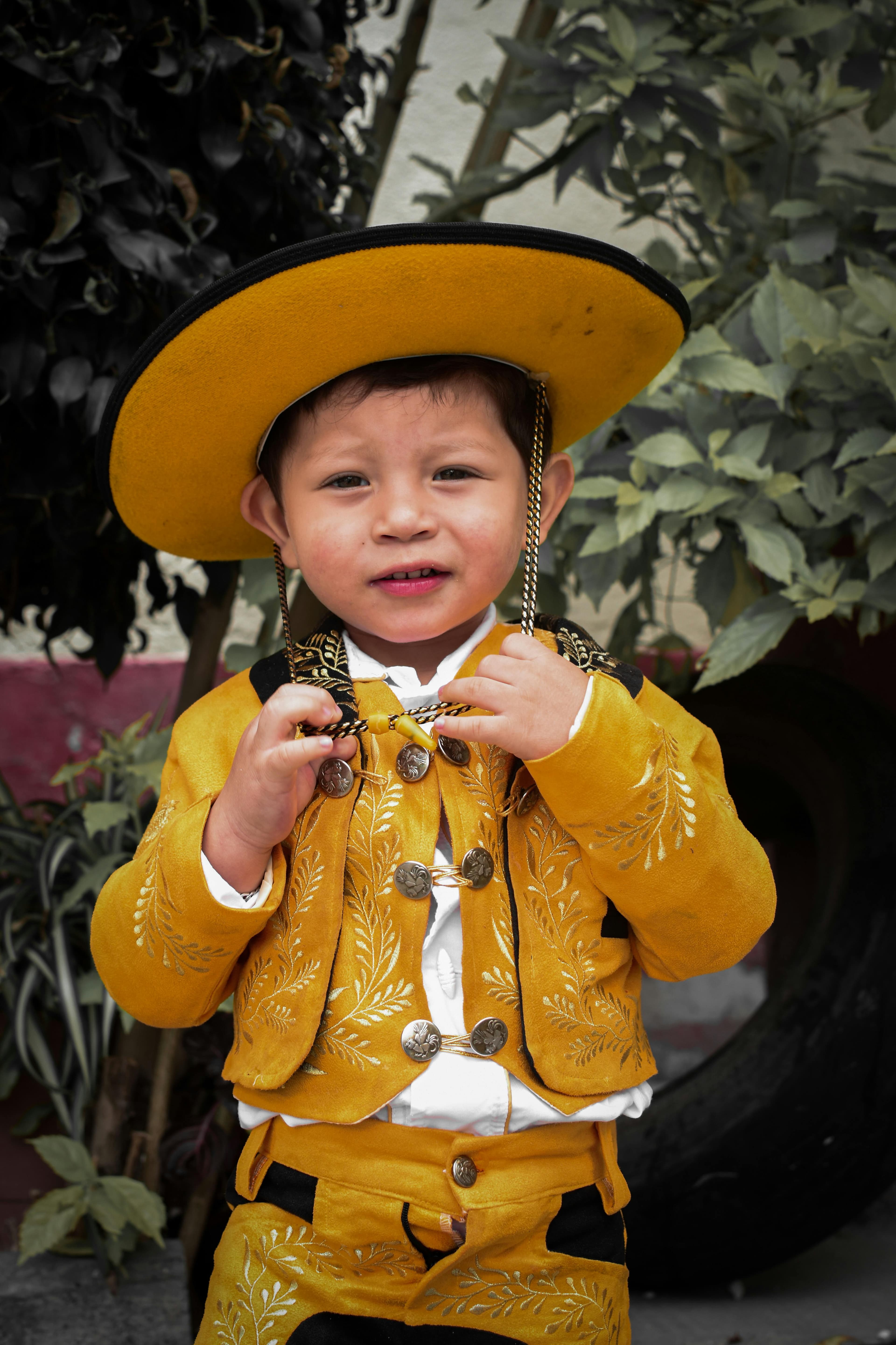 Un niño pequeño vestido con un traje tradicional amarillo y un sombrero ancho a juego, de pie frente a plantas, mirando a la cámara.