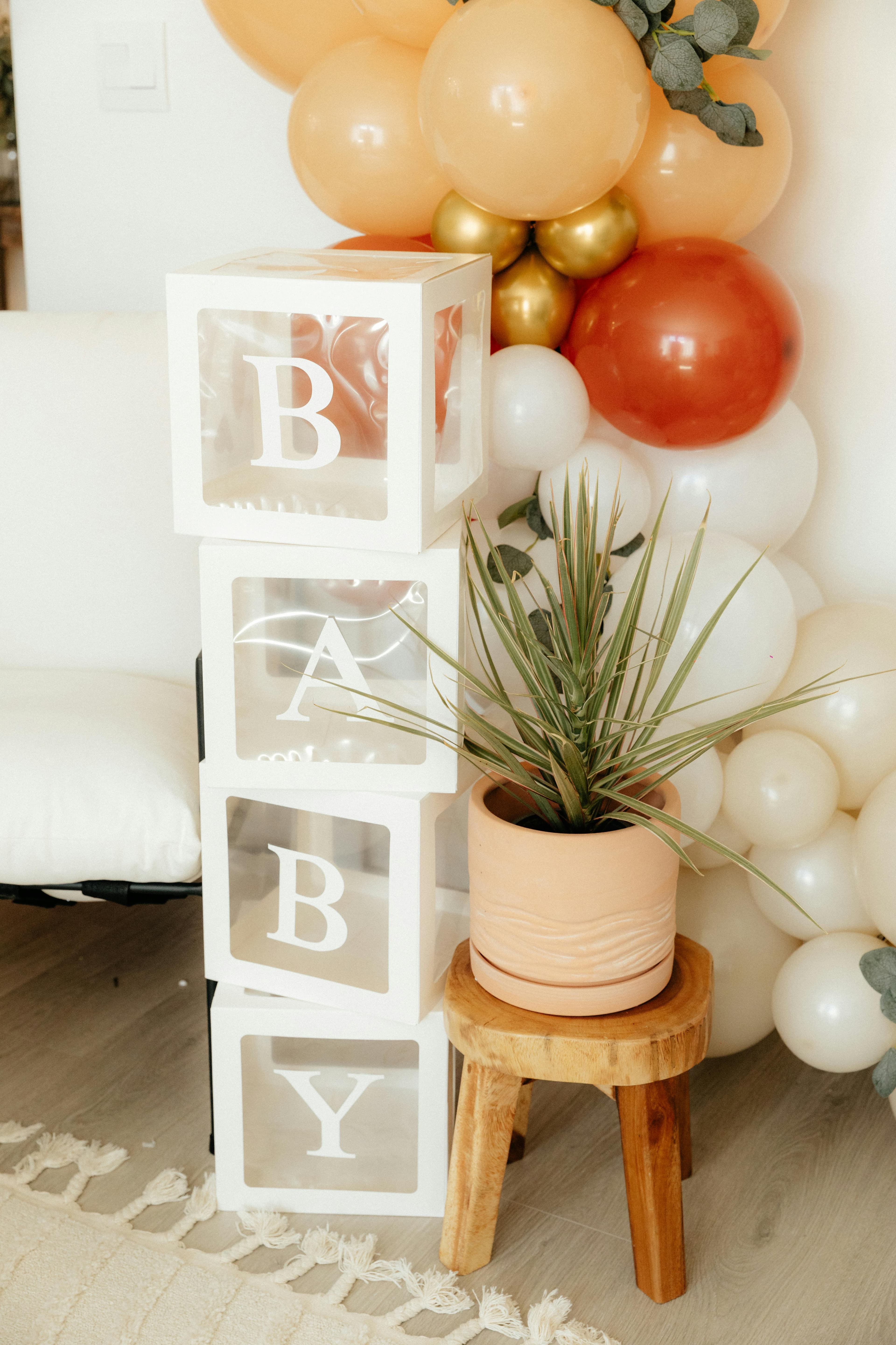 Una decoración interior con cubos transparentes apilados que forman la palabra “BABY”, junto a un arreglo de globos en tonos crema, dorado y terracota, y una planta en maceta sobre un taburete de madera.